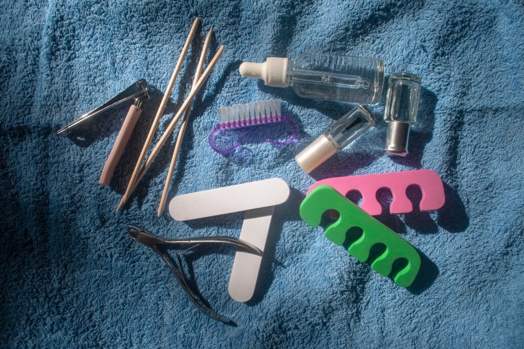Top view set of pedicure/manicure equipment on a blue towel background on a sunny day. Pedicure kit. Transparent strengthening nail polish, hand serum, clippers, nippers, nail files, nail polisher, nail brush, foam toe separators, orange wood cuticle pusher sticks. Home pedicure tools. Beauty salon, spa, nail bar, nail studio accessories.