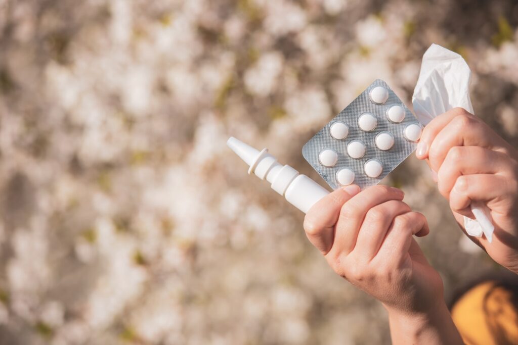 Allergy concept, young woman with pills or drugs and nose or nasal spray against the strong allergy in hand in front of blooming a tree during spring season, healthcare